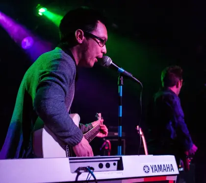 main singing in to microphone with a guitar, a keyboard in the foreground